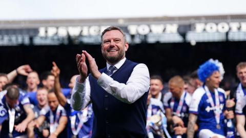 Mark Ashton is wearing a white shirt, navy blue knitted waistcoat and a striped tie. He is smiling and applauding, with Ipswich Town footballers celebrating behind him