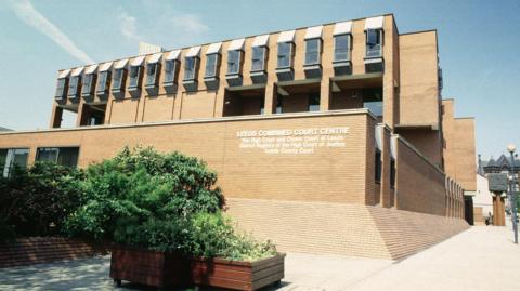Exterior of the Leeds Combined Court Centre, a modern multi-storey brick building with geometric window structures. White signage on the front wall identifies it as housing the Crown Court, County Court, and High Court District Registry.