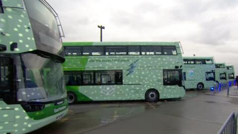 Several green double decker buses are lined up in a car park. They have a big lightening motif on the side of them with an "electric buses/Hull eah!" logo on the side of them.