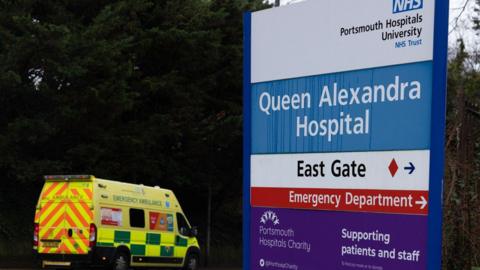 A hospital's emergency department sign with an ambulance in the background.