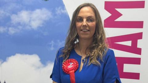 Julie Griffiths is smiling at the camera. She has long blonde wavy hair and blue eyes. She is wearing a short-sleeved blue dress with a red Labour rosette and blue heart pendant necklace. 