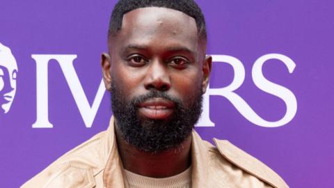 Ghetts at the  Ivor Novello Awards 2025. Image shows close-up of the rapper with a purple and white banner in the background. He has short-shaven black hair and a full short beard