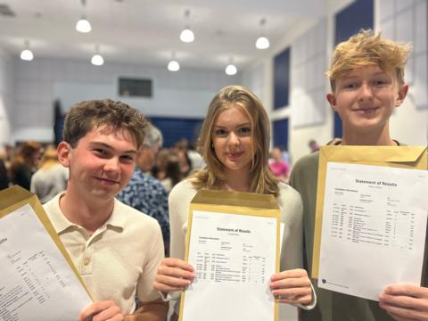 Two boys and a girl are looking at the camera and holding their GCSE results