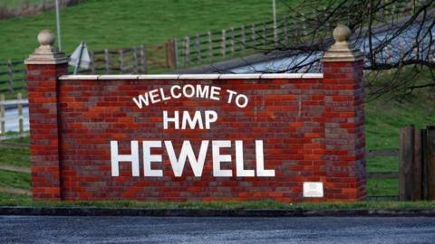 A red brick wall at the side of a road, saying 'Welcome to HMP Hewell' in white lettering. Wooden gates and grass are at the side of the road.
