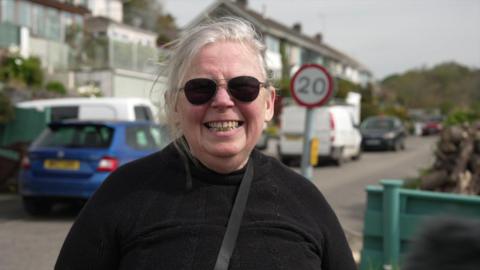 A woman standing on a street. She is wearing sunglasses and is smiling into the camera. Parked cars and houses are behind her.