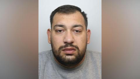 A police mugshot of a man in his twenties, with dark hair and a short beard. He is wearing a grey t-shirt and staring directly into the camera.