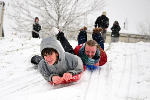 Two teenagers grin as they slide down a snowy hill towards the camera on plastic sledges