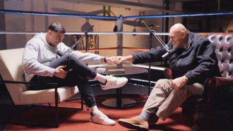 Tommy (left) and John (right) Fury sitting in arm chairs in a boxing ring, fist-pumping