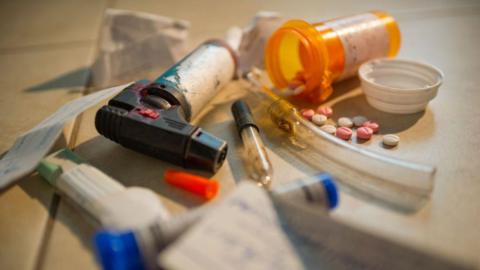 This image has multiple pieces of illegal drug equipment. There are clear tubes, a small orange bottle with multi colour pills inside, a small flame torch and small pieces of paper. 