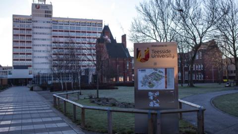 Teesside University's campus. A sign containing a map of the campus stands  on a lawn outside one of the university buildings. There are a number of trees behind it, all without leaves.