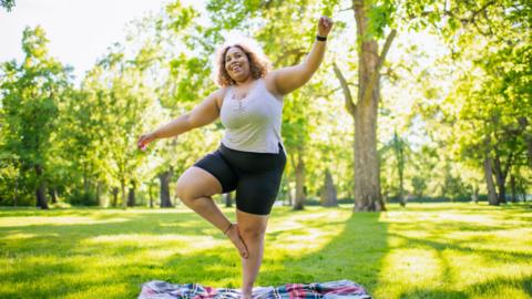 A woman stands on one leg, stood on a picnic blanket on a patch of green grass with trees in the background