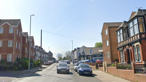 Cars are driving along the road or parked at either side. There are flats and houses or businesses to either side. Two junctions can be seen - one to the left and one further along on the right hand side.