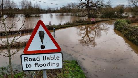 A flooded rural road and field with in the foreground a warning road sign saying "Road liable to flooding"