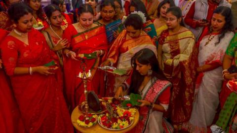 Group of ladies - dressed in indian colourful outfits standing around a candle and trays of flowers 