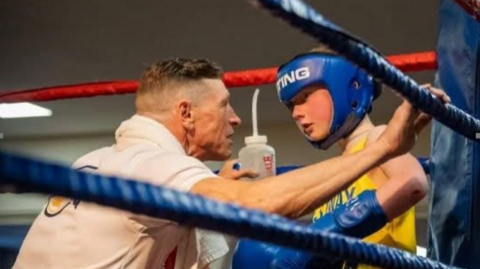 A boxing club coach, on the left, talking to a young boxer, on the right of the image. The coach is wearing a white T-shirt and has a towel around his neck. The fighter is wearing blue boxing gloves and blue protective headgear.