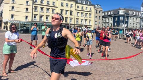 James crosses the finish line of a marathon in an urban square, triumphantly holding a golden inflatable number '7'. A red ribbon marks the finish, held by another person. Behind James, other participants cheer and clap, with tall buildings and a clear blue sky in the background