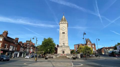 In the centre of the image of a town centre is a large light-coloured stone column memorial. It has a pointed slate roof and a clock as well as brass plaques. Near the plaques are red poppy wreaths. The memorial is on an island in the middle of a road - on either side are shops and cars.