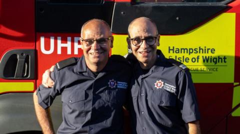 Dave and Paul Minall smiling in front of a Hampshire & Isle of Wight Fire and Rescue Service vehicle. They have their arms around each other's shoulders.
