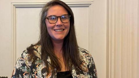 Charlotte Traynor, a woman with long dark hair, she wears large round glasses and has a bright smile, she wears a patterned white and black jacket.