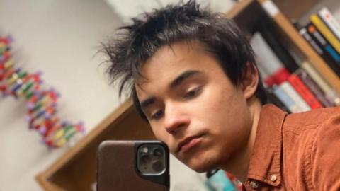 Moses Fernandes posing for a selfie photograph. He is wearing a brown corduroy shirt and is standing in a room with furniture behind him. He has medium-length black hair.