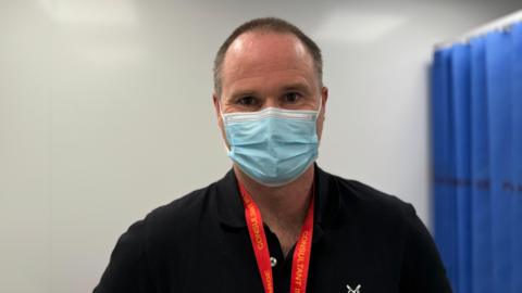 Will Niven, emergency medicine consultant, chief of service for access and medicine and at the Royal Surrey NHS Foundation Trust. Will is wearing a black shirt, mask and red lanyard.
