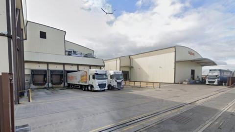 A Google street view image of white factory building, with white trucks parked next to it.