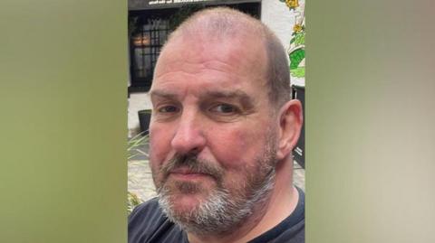 Man with grey short hair and beard in grey top smiling at camera 