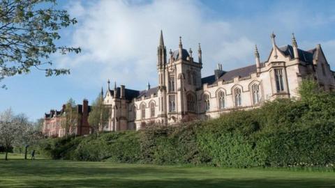 The university campus in Londonderry. A number of bushes and grassy areas can be seen in front of a Gothic-looking building with several large windows.