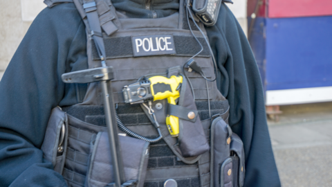 A person wearing a black top with a bullet proof vest on top with a number of pockets and a yellow gun in the middle. The word POLICE is written across the vest. 