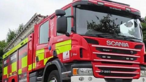 Stock image of a fire engine, with a ladder on top, that is parked in front of some trees 