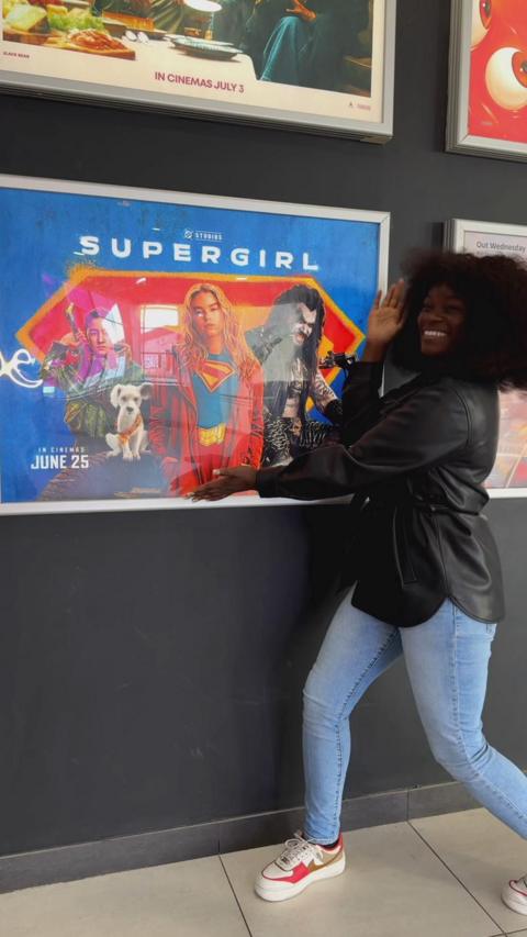 A woman in a black leather jacket is smiling at the camera. She is standing next to a Supergirl film poster