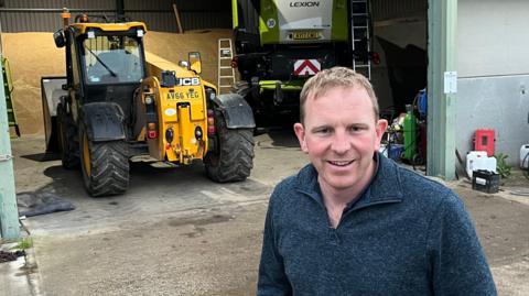 Tom Mead smiles and looks directly at the camera as he is photographed outside in front of a yellow tractor. He is wearing a blue jumper and dark trousers and has both hands in his pockets. Behind him is a large barn with farmyard items on the ground behind. 