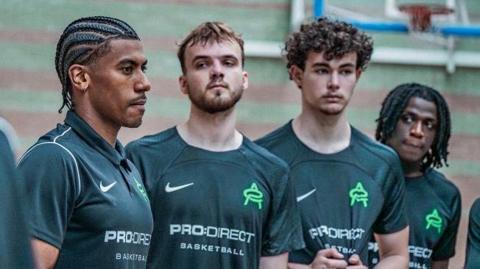 Four men in green PRO:DIRECT Basketball T-shirts. Andre Arissol on the far left has short dark hair and beard. They are standing in front of a basketball hoop.