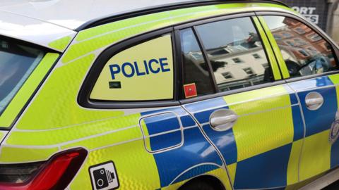 The side of a police car. It has reflective blue and yellow checks, the lettering POLICE on the back window and a sticker with the picture of a speed camera on it. Buildings are reflected in its windows.