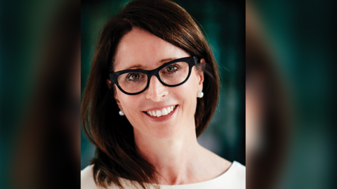 A shot of Cara looking straight into the camera, she has long dark hair, is wearing pearl-type earrings, a white top and glasses.