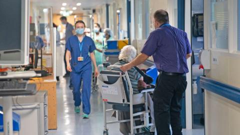 A woman in blue scrubs walks towards the camera with a face mask on, while a man, with his back to the camera, wheels an elderly person with white hair down a hospital corridor on a bed.