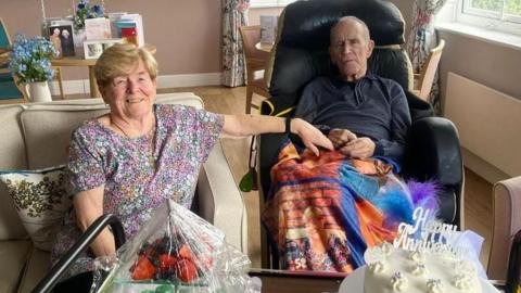 The couple at their care home. Mr Meskell sits in a wheelchair holding his wife's hand to the left. A cake with a happy anniversary sits at the front. 