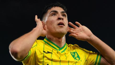 Marcelino Nunez cups his ears while wearing a yellow and green Norwich City top at night.