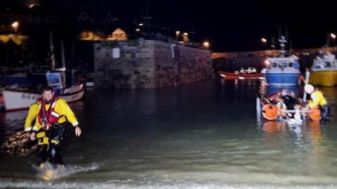 Three members of the Newquay RNLI crew wade back to shore in the town's harbour following a rescue. All three crew are wearing black and yellow overalls and red lifejackets. One of the crew is making an adjustment to an orange rib boat.