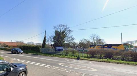 A Google image of a road junction shows part of a car driving towards a larger road, with another car on the left. There is an industrial unit in the background.