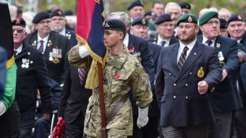 A young male soldier in Army cap and camouflage uniform jacket leads a march while holding a ceremonial standard flag. Veterans in dark suits, regimental caps and medals on lapels march behind him during the parade.