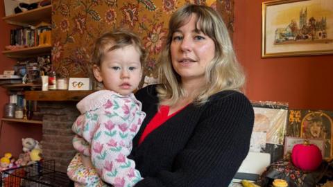 A woman with blonde hair wearing a black cardigan and red top is cradling her toddler in her arms. Her toddler is wearing a knitted cardigan with pink flowers on. They are standing in their living room which has vinyls propped up on the wall, a fireplace and a print of a church and trees on the wall. There is also a shelf full of books and board games.