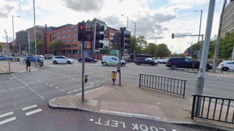 A wide road junction with traffic lights and pedestrian crossings and tall buildings on the far side