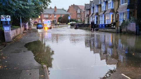 Street with terraced houses running along and some residents standing outside looking at the flooded road.