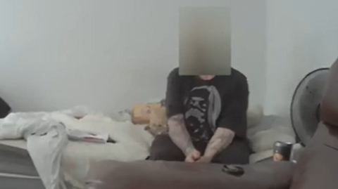 A man with a blurred face is sitting on a messy bed wearing a black T-Shirt.