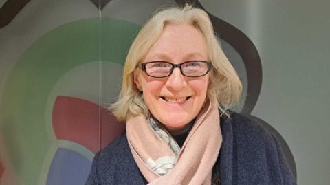 A woman with slightly shorter than shoulder length blonde hair stands wearing a grey coat and pink scarf against the backdrop of a West Northamptonshire Council logo.