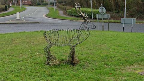 The wire-framed stag sculpture in the middle of a grass-covered roundabout with a road snaking of in the background