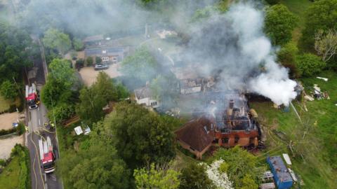 A house surrounded by trees is charred and burnt as smoke billows into the sky.