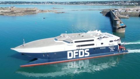 Large DFDS high-speed ferry sailing through calm turquoise water near Jersey harbour entrance, with a breakwater, port facilities, and St Aubin visible in the background under a clear sky.