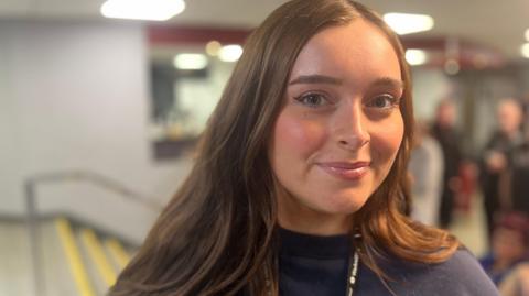 Elsie Booth has long brown hair and is smiling while wearing a navy top.